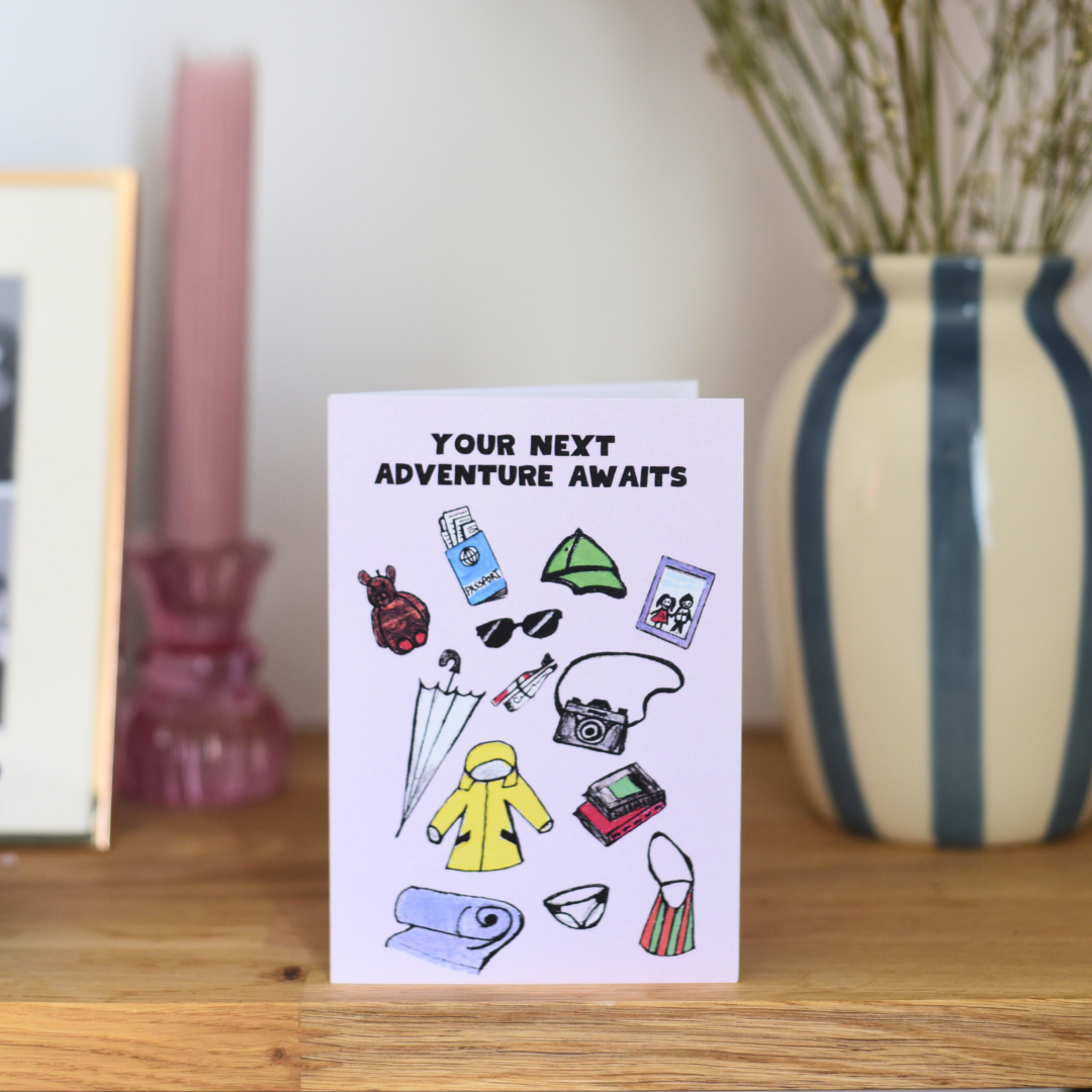 Greeting card with travel-themed illustrations and 'Your Next Adventure Awaits' text on a wooden surface.