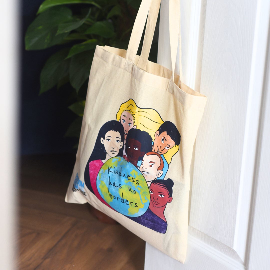 Tote bag with a colorful design of diverse people around the world, with text 'Kindness has no borders', hanging on a white door.